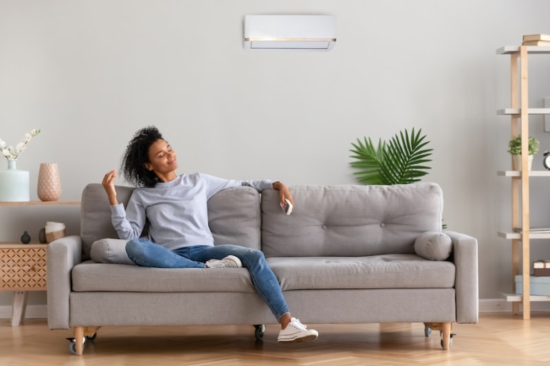 Ductless Mini Splits for Comfortable and Healthy Living - African american relaxed woman sitting on comfortable couch in living room at modern home holds air conditioner remote control enjoying breathing fresh cool air at summer or warm air at winter season (African american relaxed woman sitting on couch.