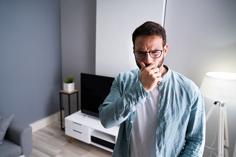 A man wearing glasses and a light blue shirt stands in a modern living room, holding a fist up to his mouth as he coughs.