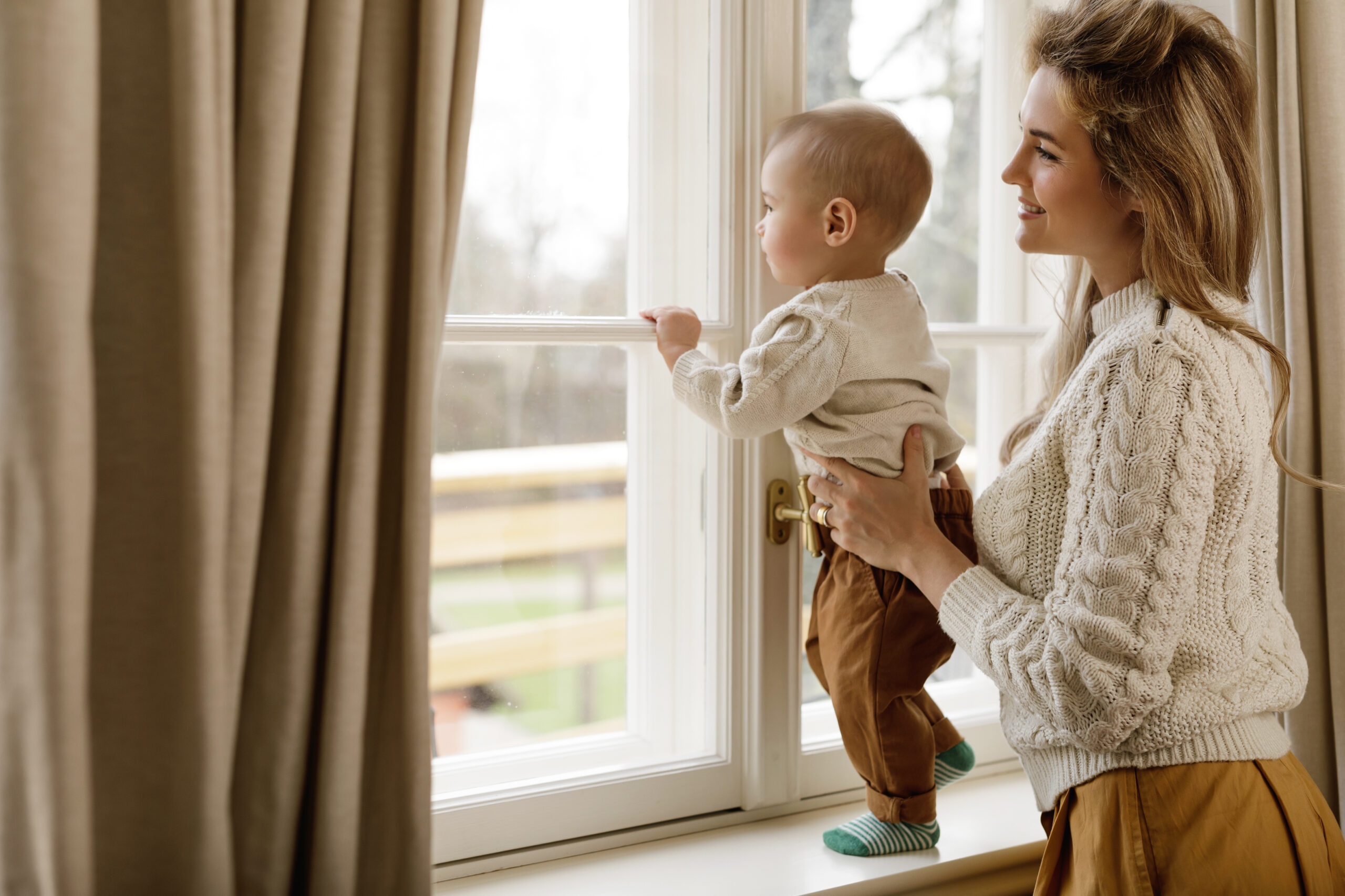 Mother and her little baby son wearing warm sweaters standing by the window